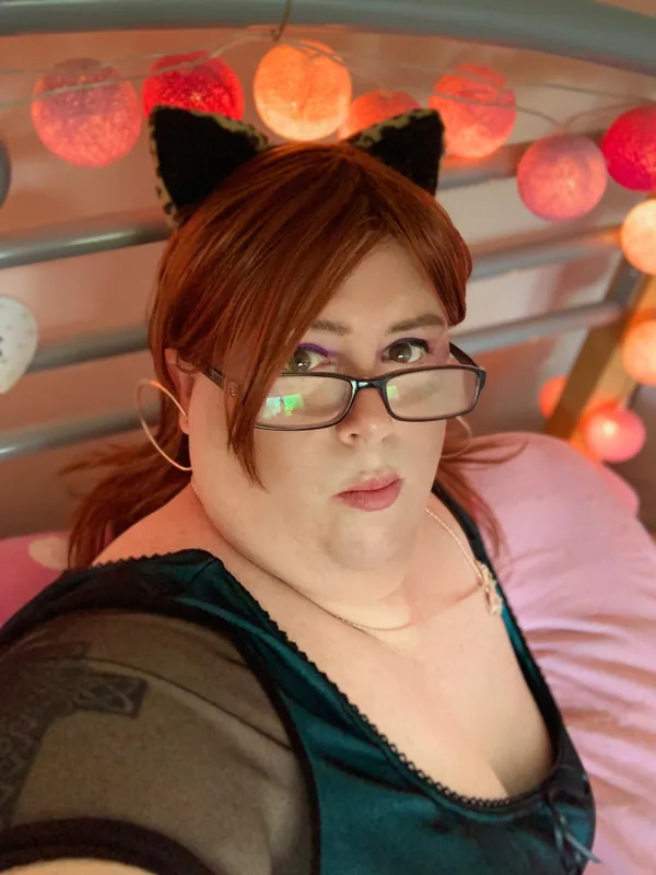 Portrait d'une femme aux cheveux châtain clair, lunettes, oreilles de chat, haut vert, sur lit avec lumières colorées.