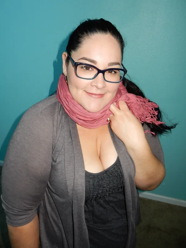Portrait en buste d'une femme aux cheveux noirs, lunettes, foulard rose et cardigan gris devant un mur turquoise.