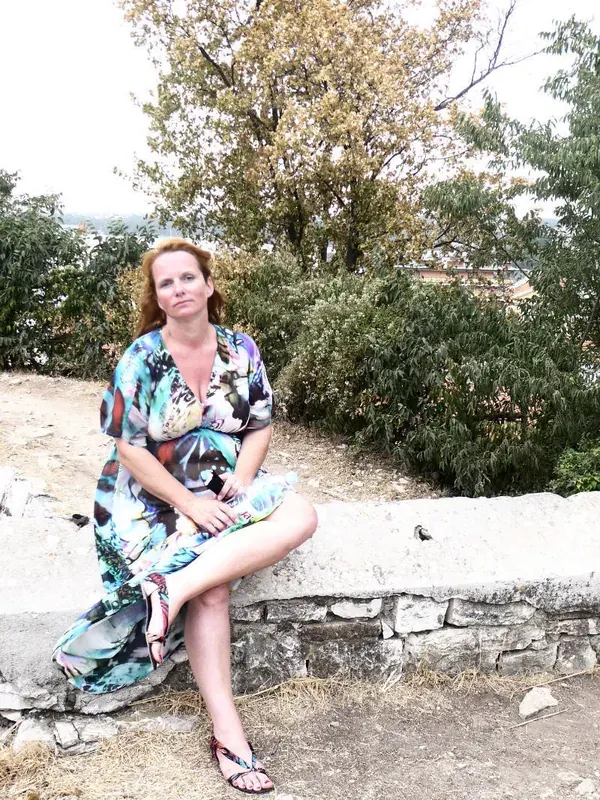 Femme aux cheveux roux assise sur un mur de pierre en robe colorée, dans un cadre naturel avec arbres et végétation.
