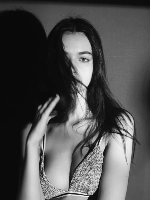 Portrait en noir et blanc d'une femme aux cheveux longs portant un soutien-gorge, un œil visible, regardant l'objectif, main près du visage.