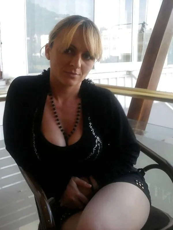 Portrait d'une femme assise près d'une fenêtre, vêtue de noir avec un décolleté profond, regardant l'objectif.