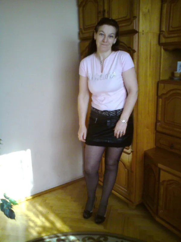Femme debout en intérieur, vêtue d'un t-shirt rose et d'une jupe noire, devant des meubles en bois.