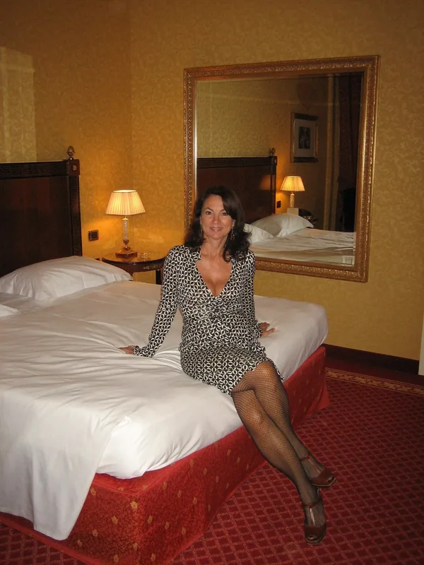 Femme assise sur un lit dans une chambre d'hôtel, portant une robe à motifs, des collants à mailles et des sandales.
