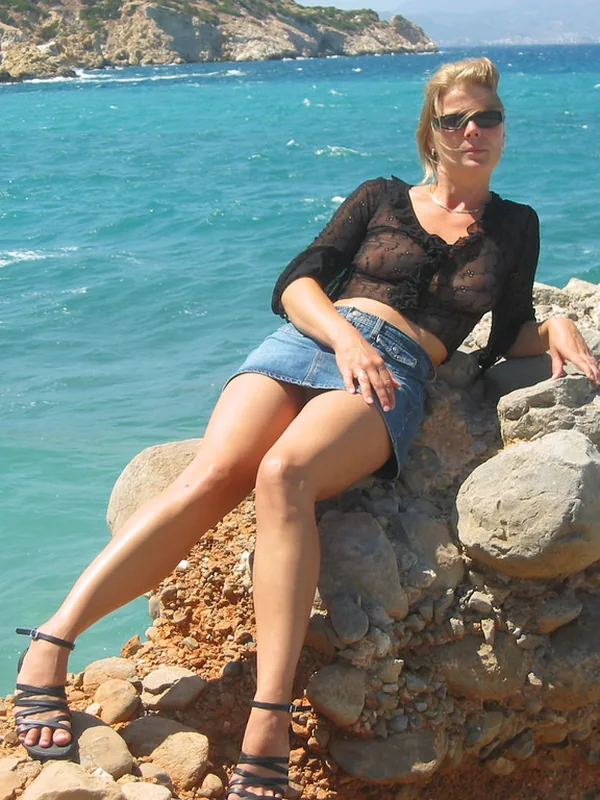 Femme blonde en tenue décontractée assise sur un mur de pierres au bord de la mer turquoise.