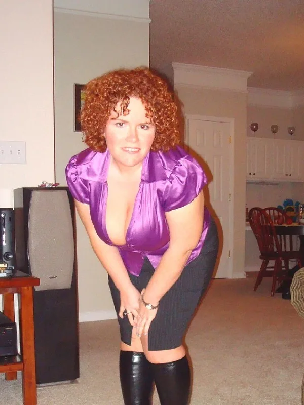 Femme aux cheveux roux bouclés portant une chemise violette, une jupe noire et des bottes, penchée en avant dans un salon.