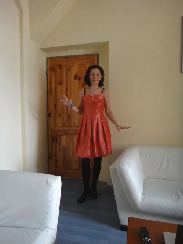 Femme en robe rouge orangé et collants noirs debout dans un salon avec canapés blancs et porte en bois.