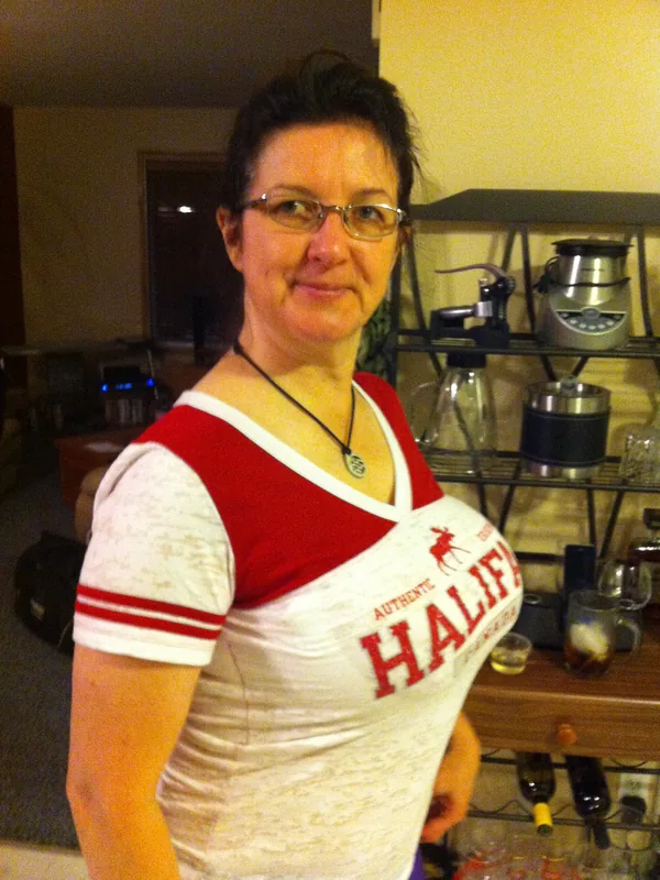 Portrait d'une femme aux cheveux courts et lunettes, portant un t-shirt blanc et rouge, dans un intérieur avec étagère de cuisine.