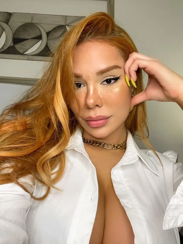 Portrait d'une femme aux cheveux cuivrés portant une chemise blanche ouverte et un collier doré, regardant l'objectif.