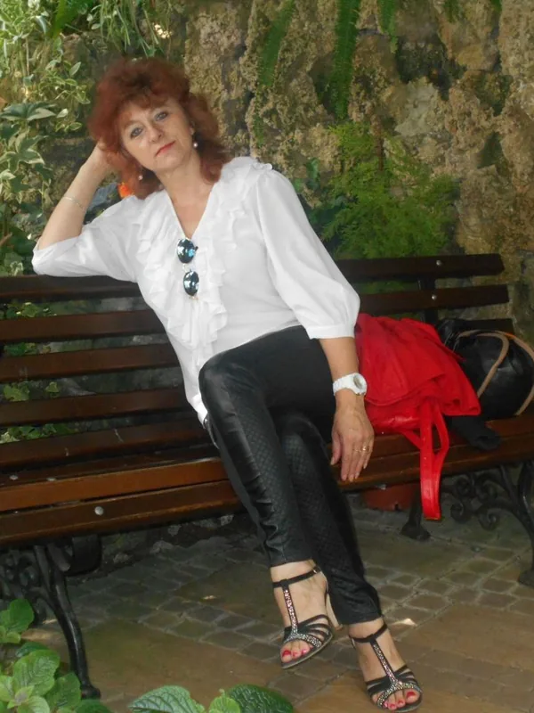 Femme aux cheveux roux assise sur un banc en bois dans un jardin, entourée de plantes vertes.